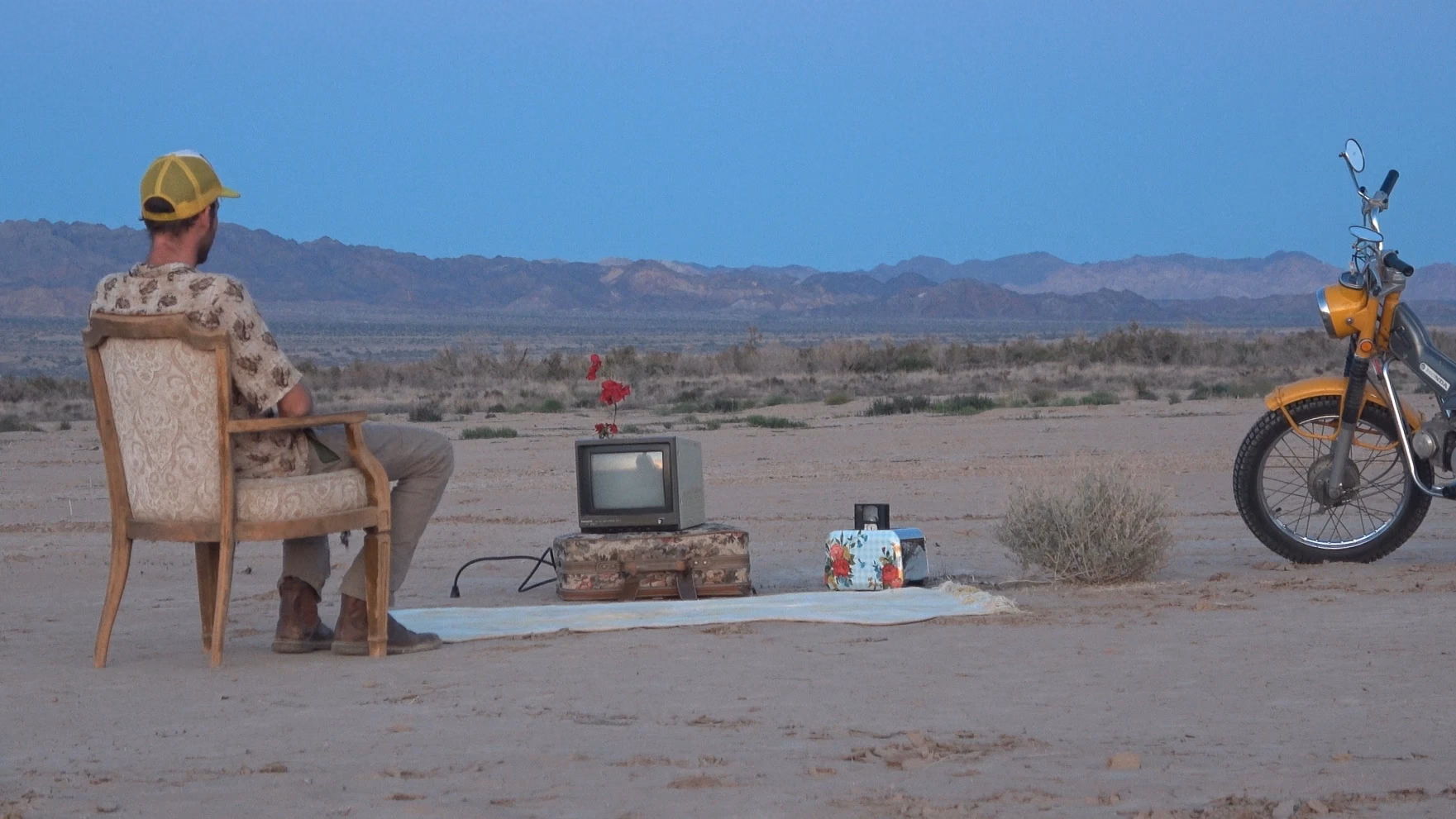 A seated figure, a television and a motorcycle in the desert